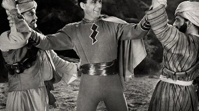 black and white image of man dressed as a superhero smiling, with two people on either side