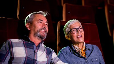 A man sitting on left and woman on right, both sitting at the cinema looking past the camera