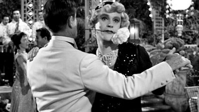 Black and white still of two men dancing, one dressed as a woman with a flower in his mouth