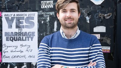 James, Queerberra, photography by Jane Duong and produced by Victoria Firth-Smith A man with brown hair and beard stands in front of a YES for Marriage Equality poster