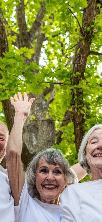 6 people in a semi circle, wearing white, smiling and raising hands against green  leaves and trees