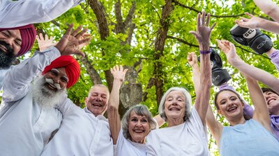 6 people in a semi circle, wearing white, smiling and raising hands against green  leaves and trees