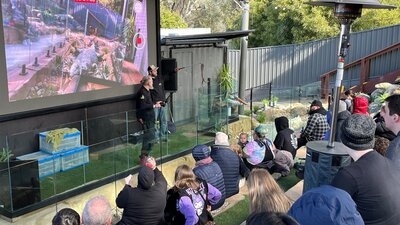 Two presenters stand on a stage with snakes while the audience watches from amphitheatre seats.