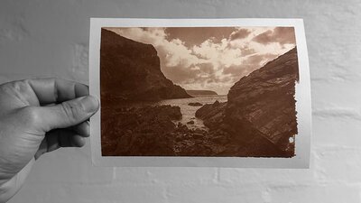 An image of a hand holding a brown-toned photograph of a rocky shore leading out to water