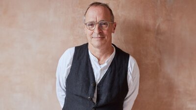 Man smiles at camera wearing white long sleeve with dark vest and glasses