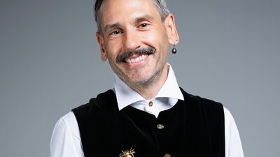A person wearing a white button up, black vest, a bee broach and a dark opal earring poses smiling.