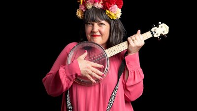 Woman holding a banjolele. She is wearing a pink dress and has a flower crown on her head.