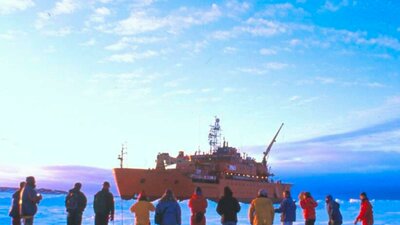 People in colorful winter gear stand on a vast icy plain, facing a large orange icebreaker ship.