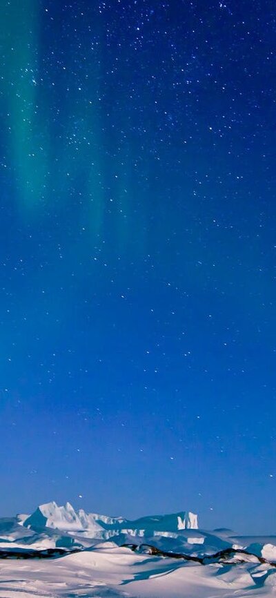 Aurora Australis and grounded icebergs near Mawson station. © Christopher (Chris) Wilson / AAD Aurora Australis glows in the night sky above a snowy, mountainous landscape.