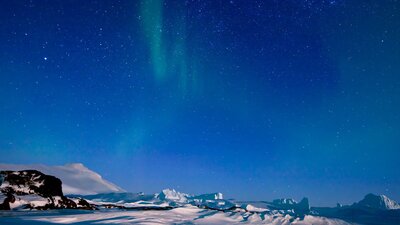 Aurora Australis and grounded icebergs near Mawson station. © Christopher (Chris) Wilson / AAD Aurora Australis glows in the night sky above a snowy, mountainous landscape.