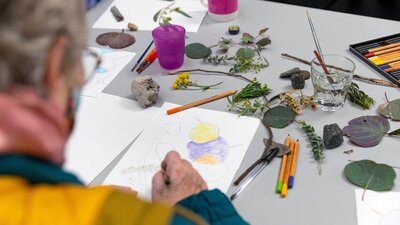 Art with Friends, 2022. National Gallery of Australia, Kamberri/Canberra. A photograph over the shoulder of an elderly person drawing gum leaves from real ones on the table