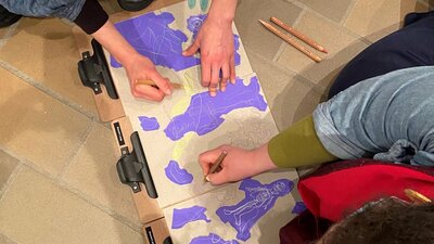 An overhead photograph of two people drawing with pencils on brown and purple paper