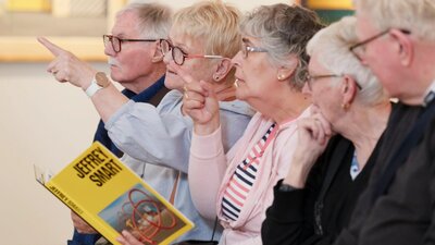 A photograph of 5 people in a gallery space pointing and looking forward