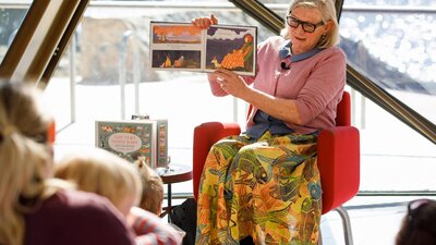 Woman sitting on a chair holding a book reading to a group of children.