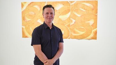 Andrew Totman stands in front of one of his artworks, a print of bright, cascading ovals or links.