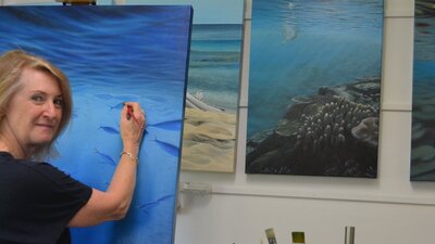 Jeanette Muirhead paints an underwater seascape in her studio, with paintings, and brushes nearby.