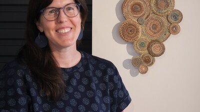 Sally Holliday stands beside her woven art handing on the wall behind her.