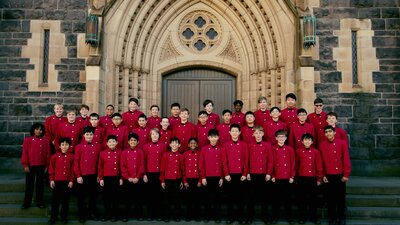 Boys choir dressed in red coats stand in front of bluestone church