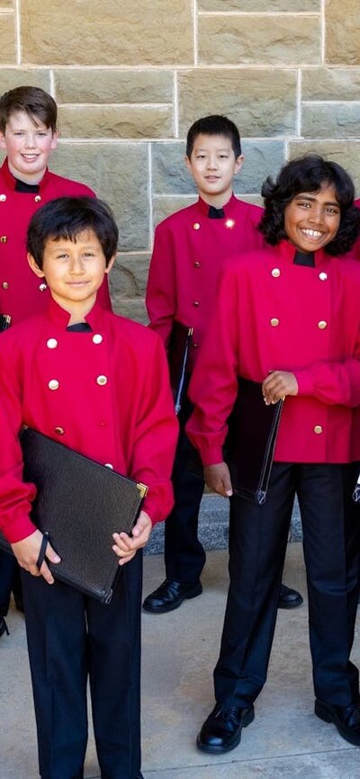 Members of boys choir stand together dressed in red coats and holding black music folders