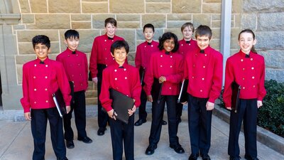 Members of boys choir stand together dressed in red coats and holding black music folders