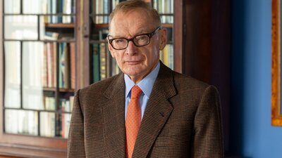 A man wearing glasses in a brown suit, standing in front of a bookcase