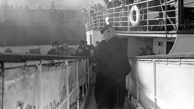 Figure standing with white cover on his head, many people surrounding the figure and watching