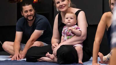 A photograph of 3 adults sitting on the floor, one is holding an infant