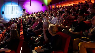 A photograph of a seated theatre audience talking with coloured lights in the background