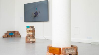 stacked glass and red bricks. TV instillation in the background featuring the artist lifting bricks.