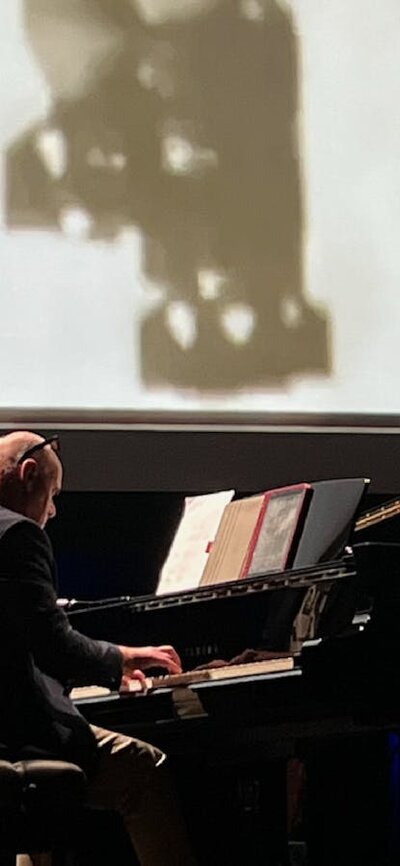 Simon Reich playing grand piano in front of screen showing bombs being dropped during World War 2