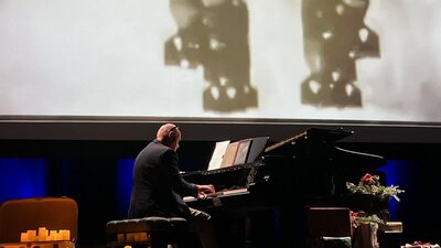 Simon Reich playing grand piano in front of screen showing bombs being dropped during World War 2
