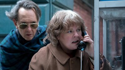 Woman on payphone looking worried, man behind her wearing sunglasses