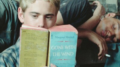 One male lying on couch sleeping, another sitting in front reading a book