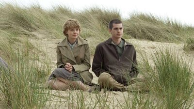 One woman and one man sitting rugged up in coats on a beach