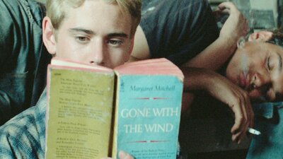 One male lying on couch sleeping, another sitting in front reading a book