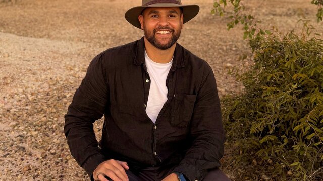 James Saunders A smiling man with a beard and a wide-brimmed hat sits on a gravelly path in a dry, bushy landscape.