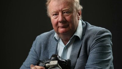 A man with grey hair wearing a blue shirt and blazer, holding a large black camera.