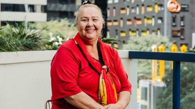 Isabel Goolagong A smiling woman sits on a balcony, wearing a red shirt and a red and yellow scarf.