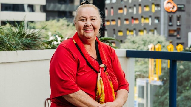 Isabel Goolagong A smiling woman sits on a balcony, wearing a red shirt and a red and yellow scarf.