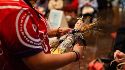 Close up of person's arms who is weaving a doll figure made from colourful wool and feathers.