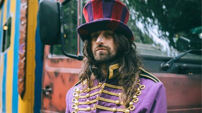 Cam Cole in a top hat and tailcoat that is purple and gold accents in front of a stripped truck.