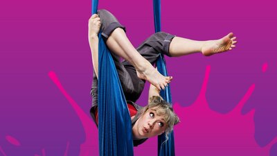 A young woman in overalls and a red shirt hangs from a coloured-blue aerial silk. Purple background.