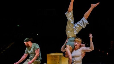 two women engage in a complex shoulder-to shoulder balance near a man laying block pathways