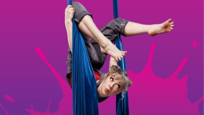A young woman in overalls and a red shirt hangs from a coloured-blue aerial silk. Purple background.