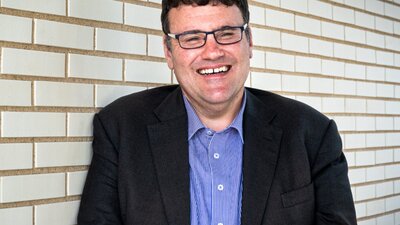 A man with glasses wearing a sttriped button down shirt and black blazer, standing in front of a whi
