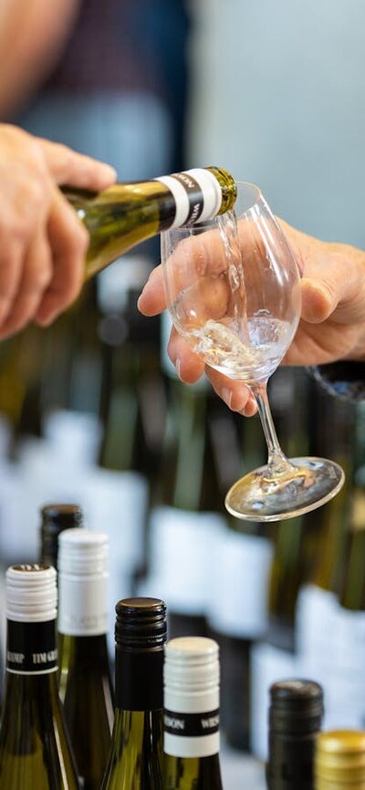 a person holding out a wine glass to have wine poured into it the background shows wine bottles