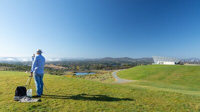 Artist painting the Canberra vista from the National Arboretum an artists easel with an artwork in front of a view of canberra and lake burley griffin