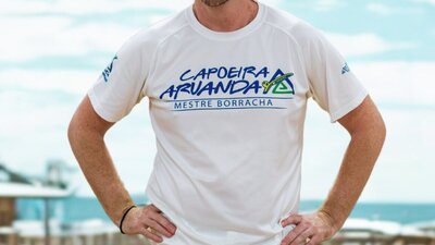 A man stands facing the camera with his hands on his hips, smiling, wearing a Capoeira tshirt