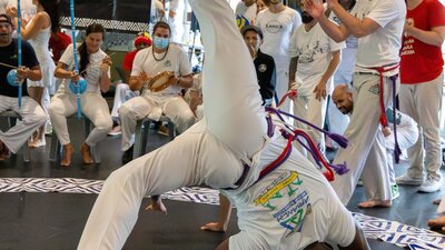 A man is mid cartwheel, playing Capoeira. There are a circle of people around him.
