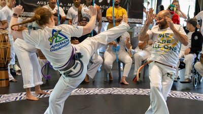 A woman and man play Capoeira, she is mid spinning kick, he is protecting his face